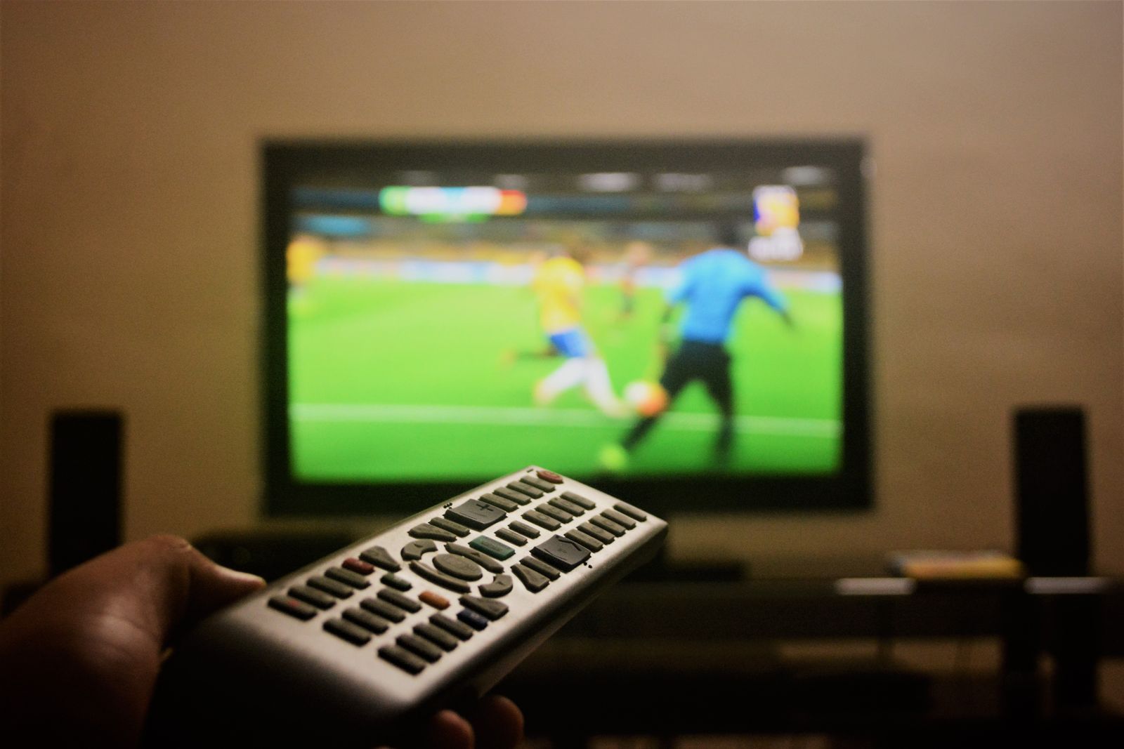 Mano con un mando a distancia frente a un televisor con un partido de fútbol