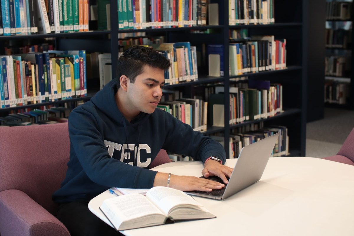 Estudiante escribiendo en un portátil junto a un libro abierto en una biblioteca